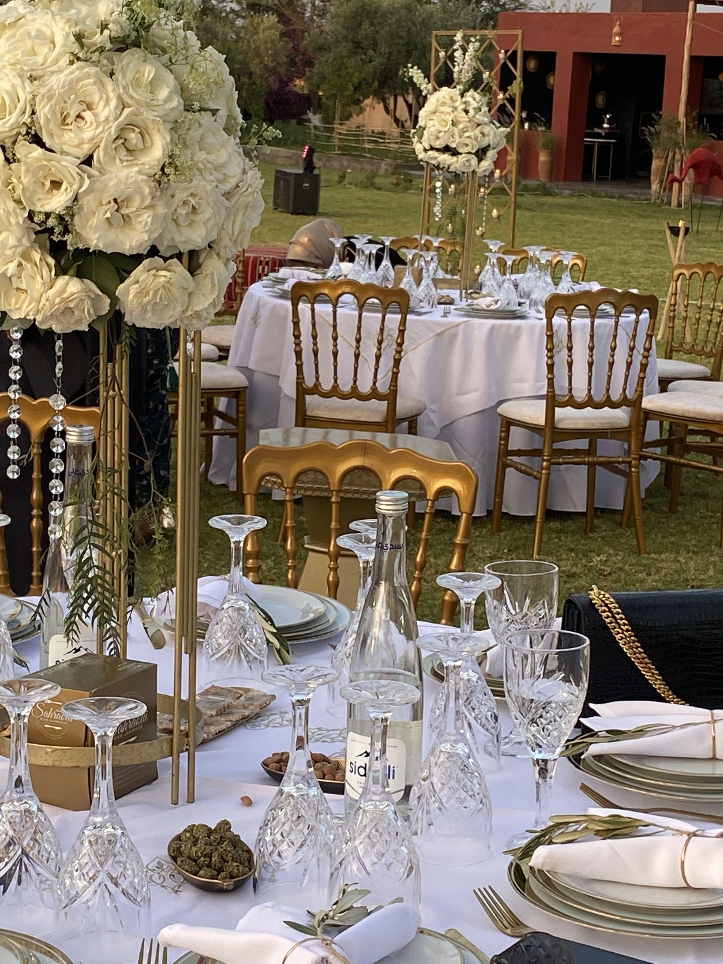 A table set for a formal outdoor event with white tablecloths, gold chairs, and crystal glassware.