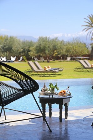 A poolside area with lounge chairs, a table with a drink and a glass, and a view of a green landscape with trees and mountains in the background.