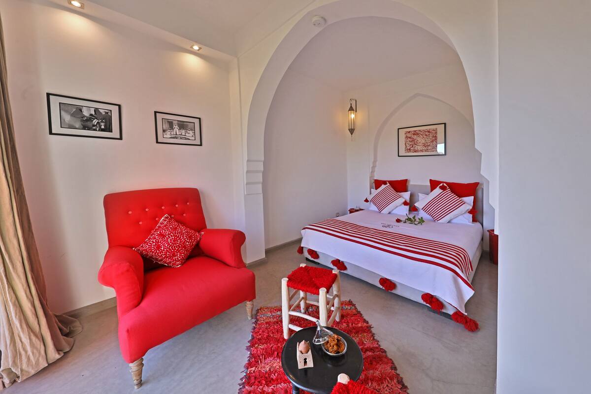 A bedroom with a red armchair, a bed with red and white bedding, and a small table with a red rug.