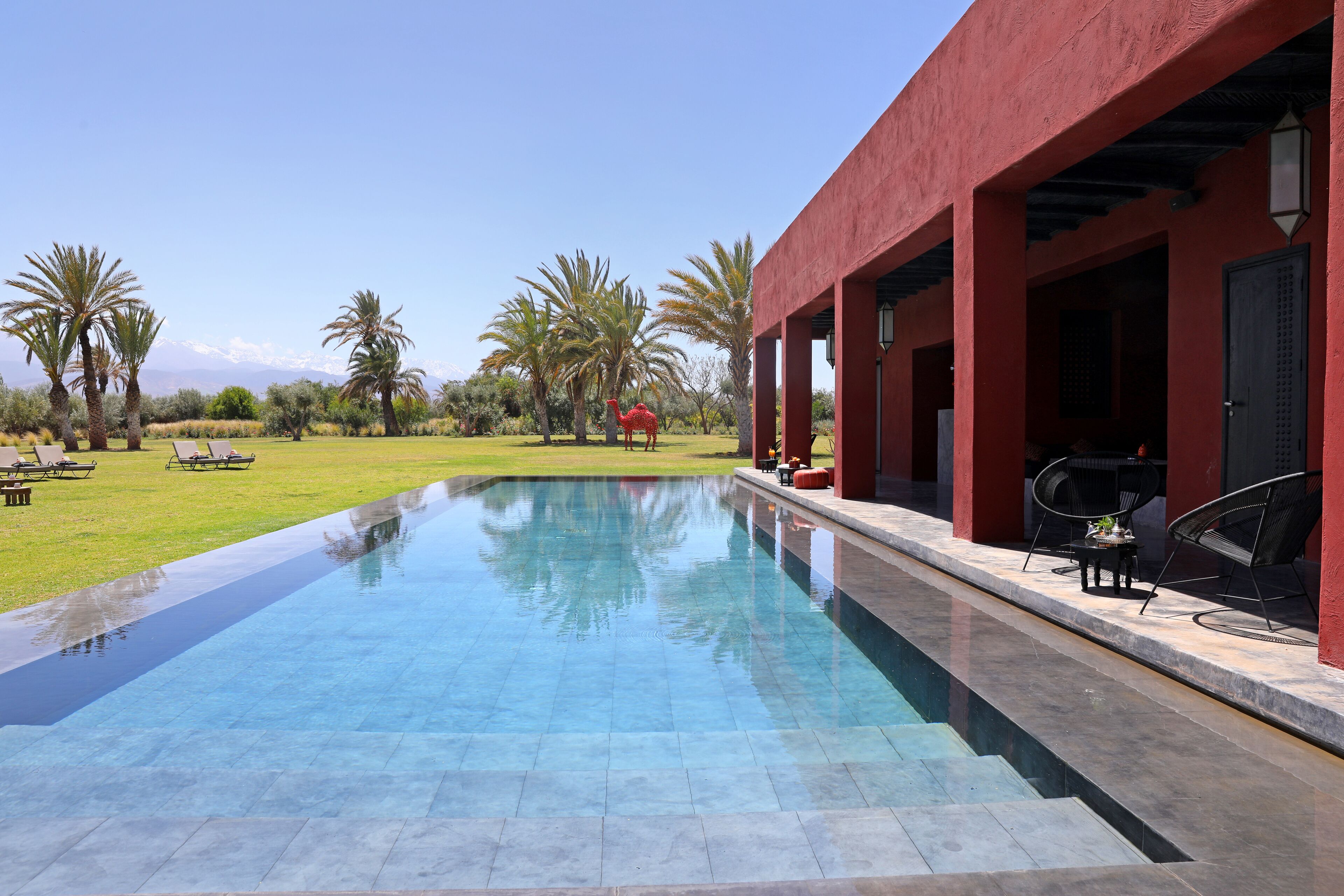 Una piscina con superficie de agua azul cristalina, rodeada por un edificio rojo con un porche cubierto.