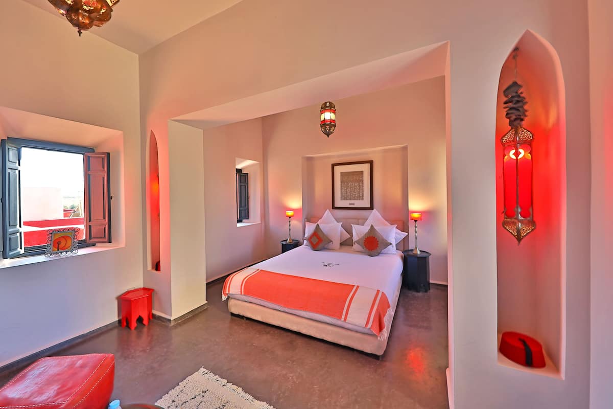 A modern bedroom with a large bed, bedside lamps, a window with a view, and a red accent wall.