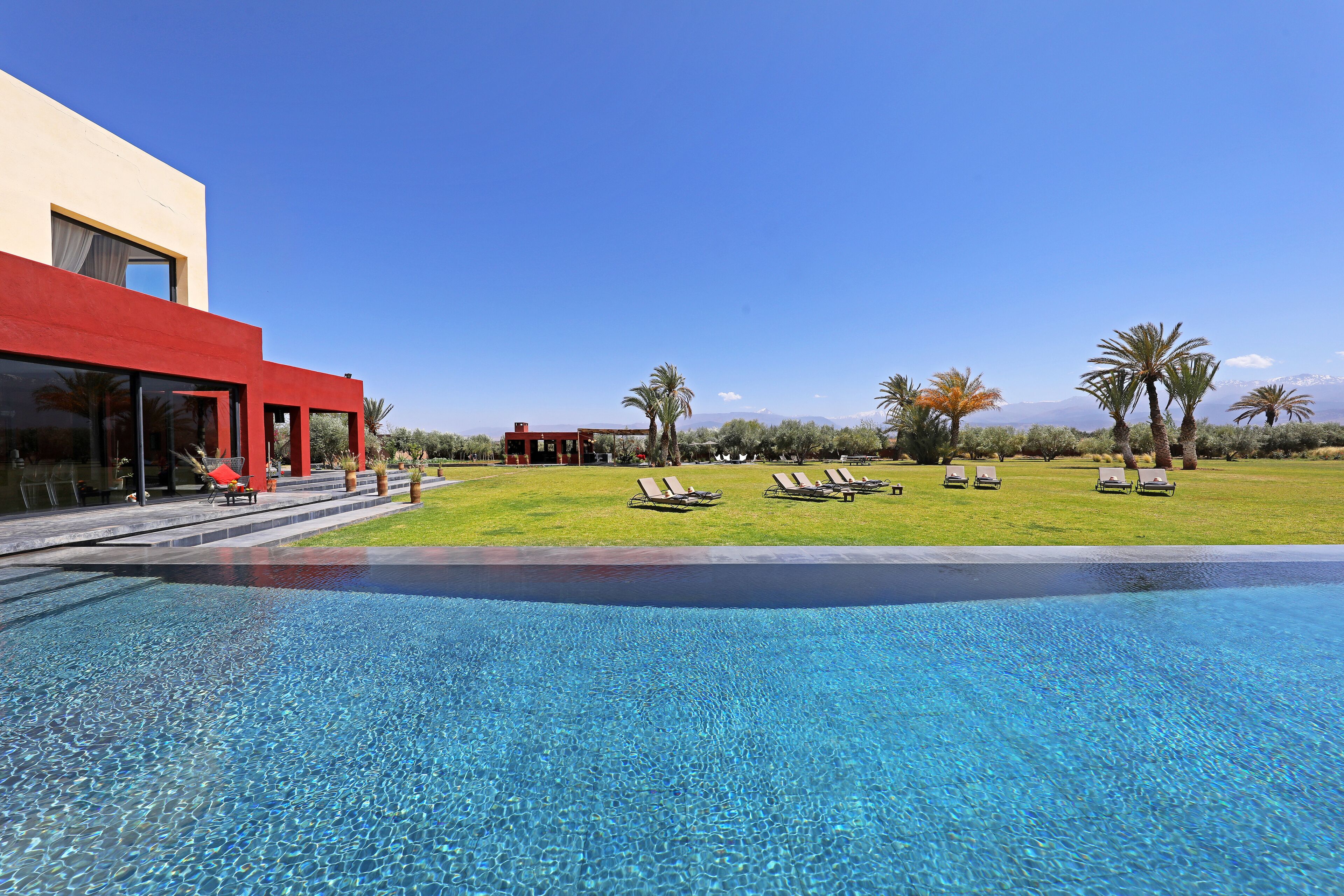 Una piscina con sillones de descanso, un edificio rojo y un área verde con palmeras.