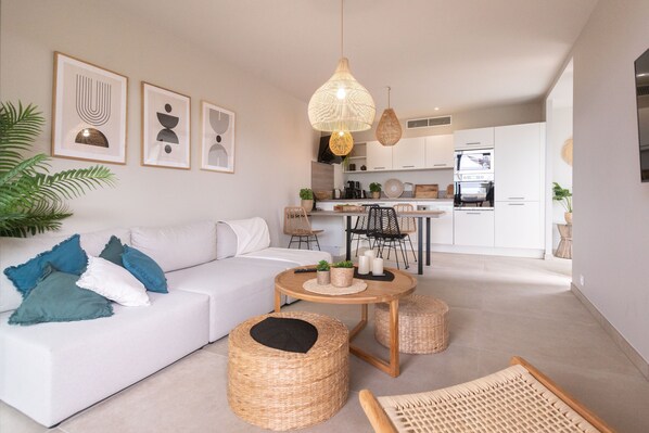 A modern living room with a white sofa, wicker ottomans, a round wooden coffee table, and a kitchen in the background.