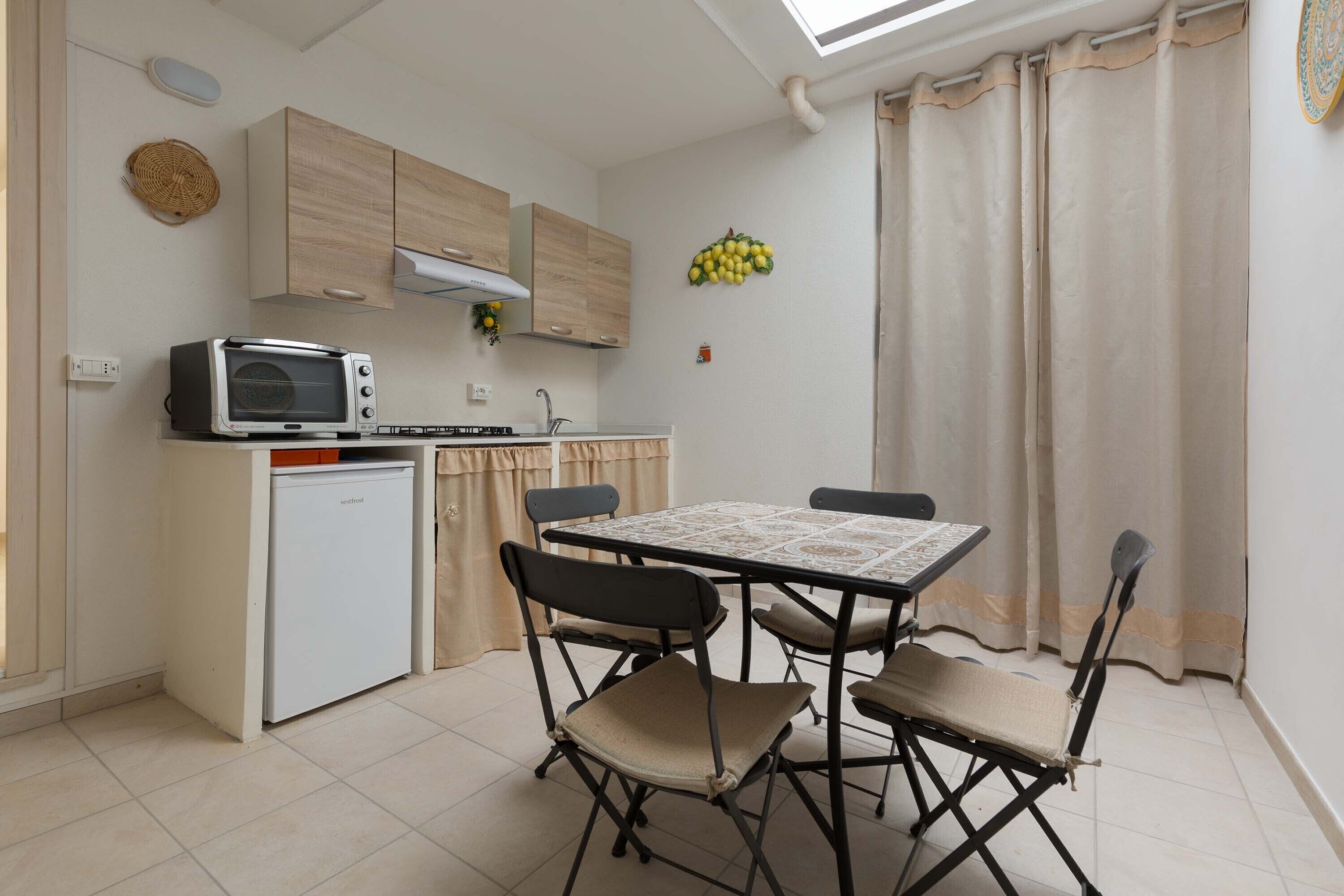 A compact kitchen with a microwave, toaster, and small refrigerator. There is a dining table with four chairs and a ceiling fan.