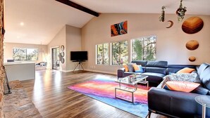 A spacious living area with a large black leather sofa, a glass coffee table, and a colorful rug. There are large windows providing natural light and a view of the outdoors.