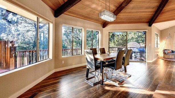 A spacious dining area with a wooden table and chairs, large windows, and a wooden ceiling.