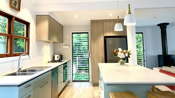 Une cuisine moderne dotée d’un îlot central, d’appareils électroménagers intégrés et offrant une vue sur la verdure grâce à une porte vitrée.