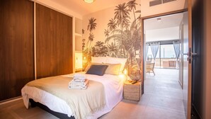 Een moderne slaapkamer met een groot bed, een houten garderobe, een muurschildering van palmbomen en uitzicht op het balkon.