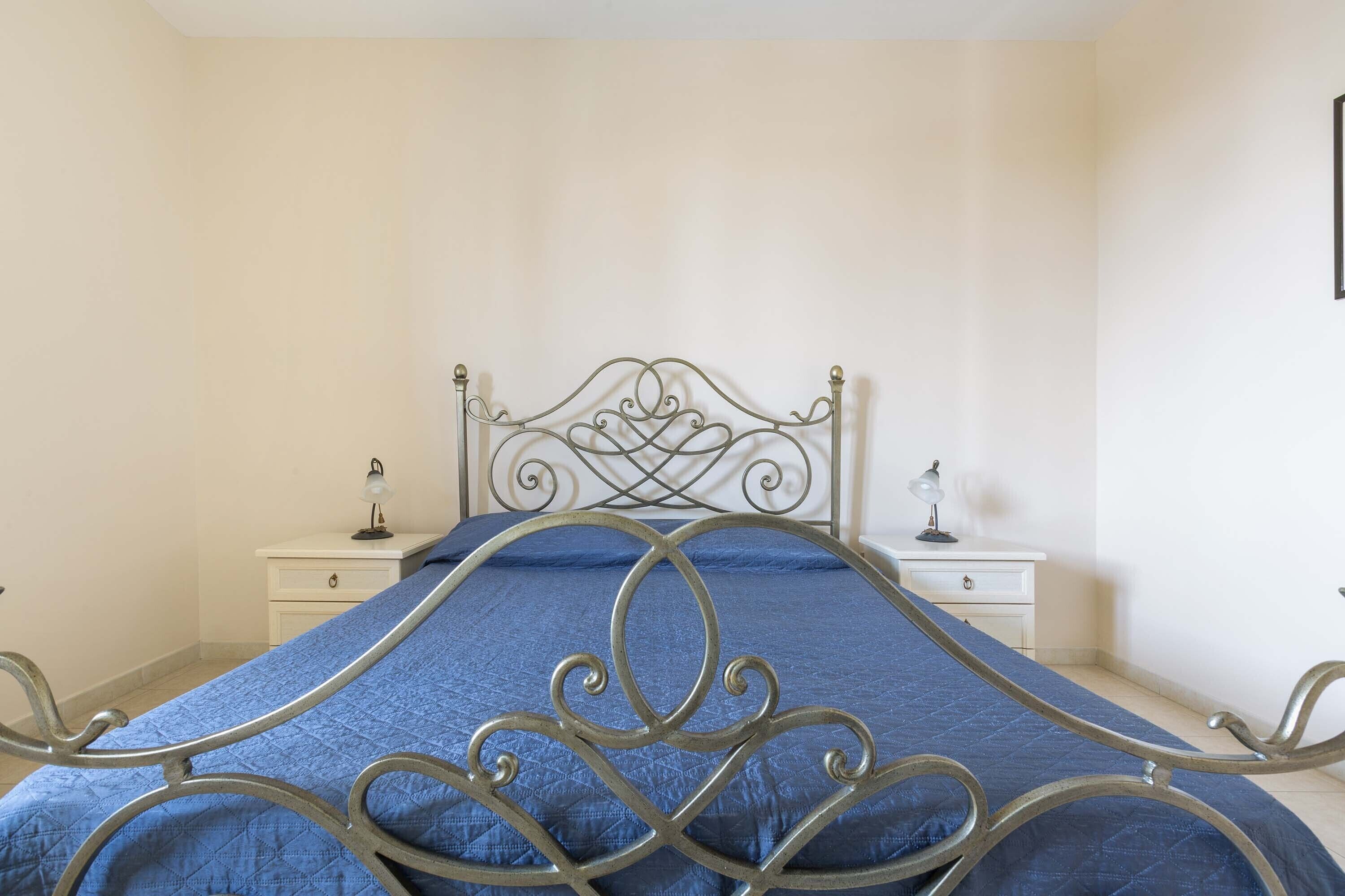 A bed with a metal frame and a blue quilt, two bedside tables with lamps, and a white wall.