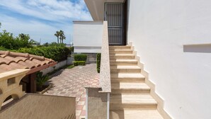 A modern building with a staircase leading to a door, surrounded by greenery and a tiled pathway. - 3010 Appartamento Kalos by Barbarhouse (Melendugno)