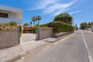 Rua residencial com uma casa moderna de dois andares, portão fechado e céu claro.