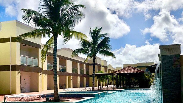 A swimming pool with palm trees, a water fountain, and a building with balconies.