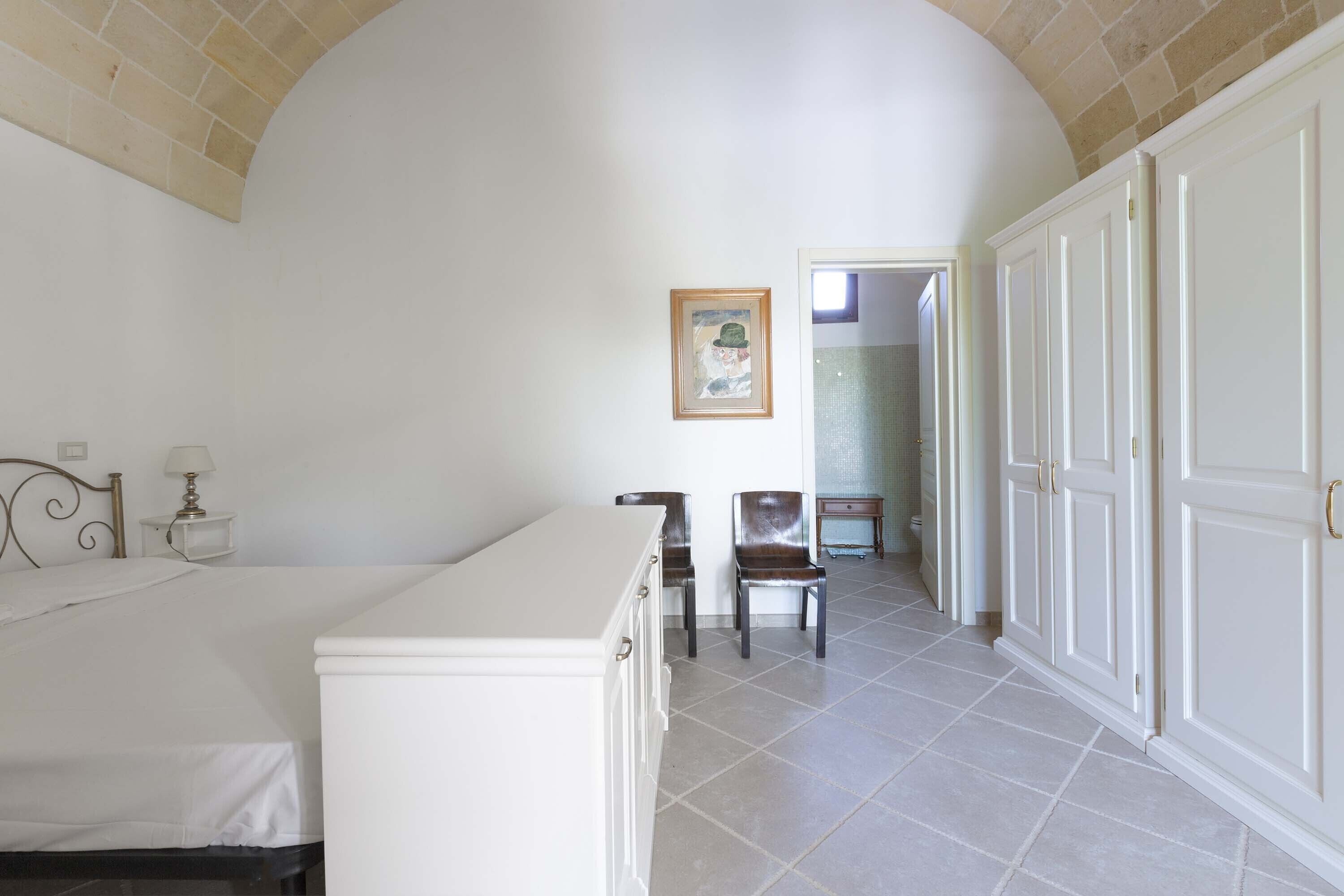 Een slaapkamer met een bed, een bureau, twee stoelen, een kledingkast en een lijst met een afbeelding aan de muur.