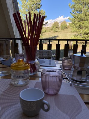 A table with a glass jar labeled "ZUCCHERO," a pink glass cup, a gray mug, and a metal container on a balcony with a view of trees and mountains.
