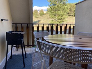A balcony with a round table and two chairs, a black metal rack, and a view of a grassy area with trees and a fence.