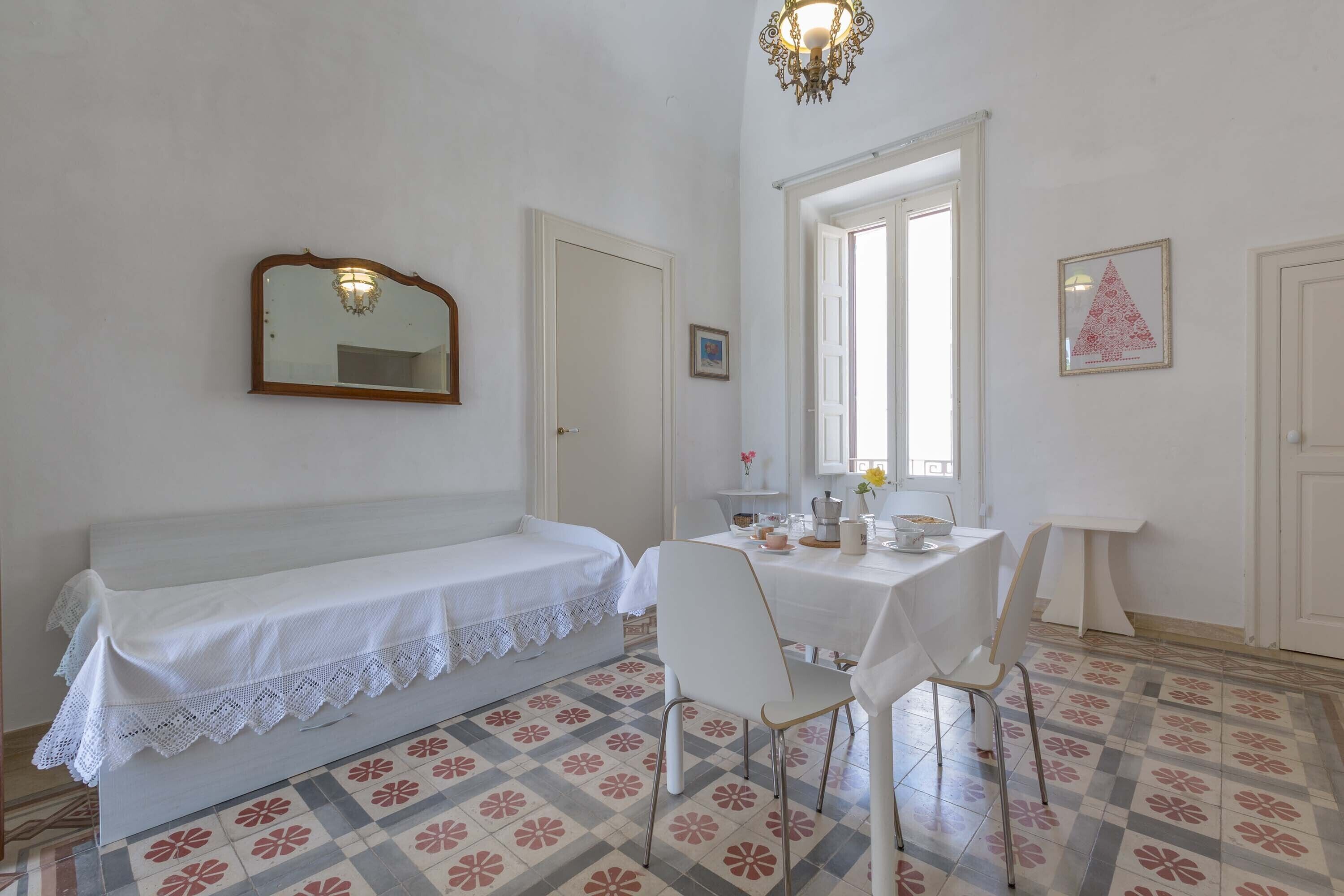 A room with a white bench, a table with chairs, a chandelier, and a patterned floor.