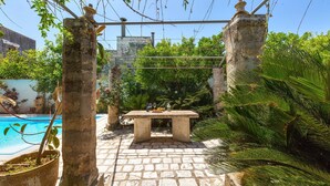 Un patio de piedra con una mesa y sillas, rodeado de abundante vegetación y una piscina.