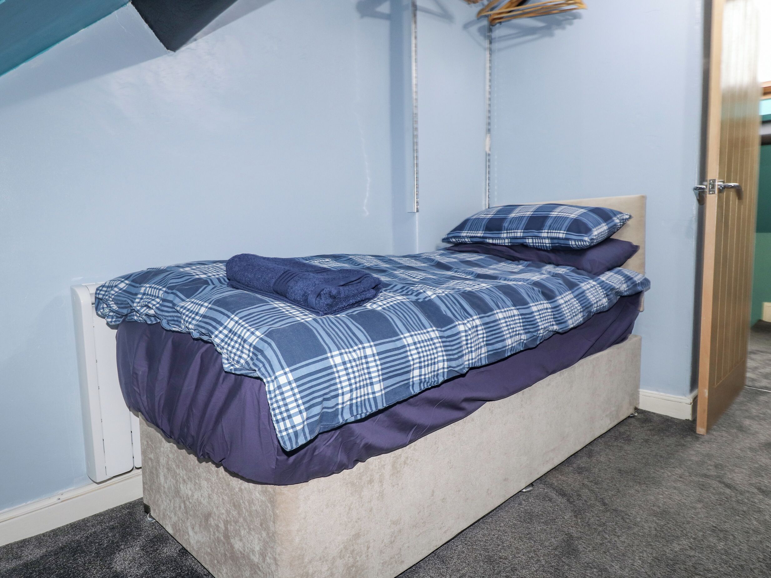 A single bed with a blue and white checkered bedspread, a matching pillow, and a folded towel on top. The bed is positioned against a light blue wall with a wooden door to the right.