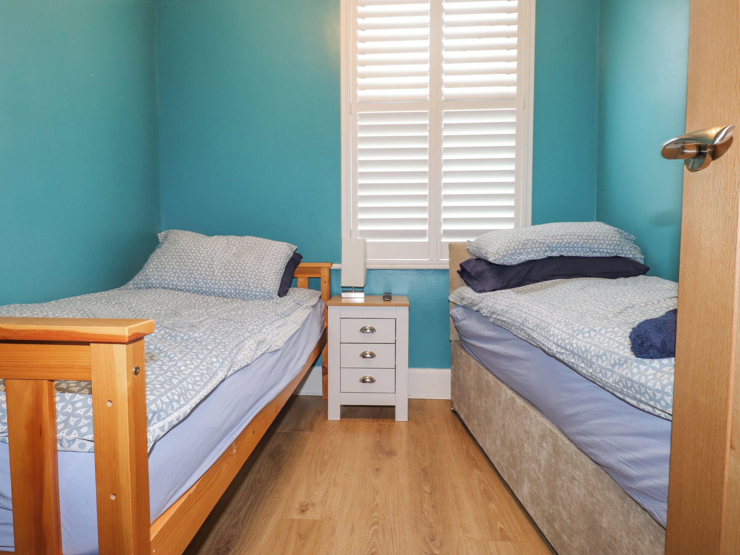 Une petite chambre à coucher, de taille réduite, avec deux lits simples, une table de chevet et une fenêtre avec des stores.