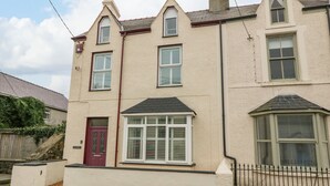 Une maison à deux étages avec une porte rouge, de nombreuses fenêtres avec volets et un petit espace clos à l’avant.