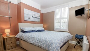 Une chambre à coucher avec un lit, une table de chevet avec une lampe, une fenêtre avec des stores et un téléviseur fixé au mur.
