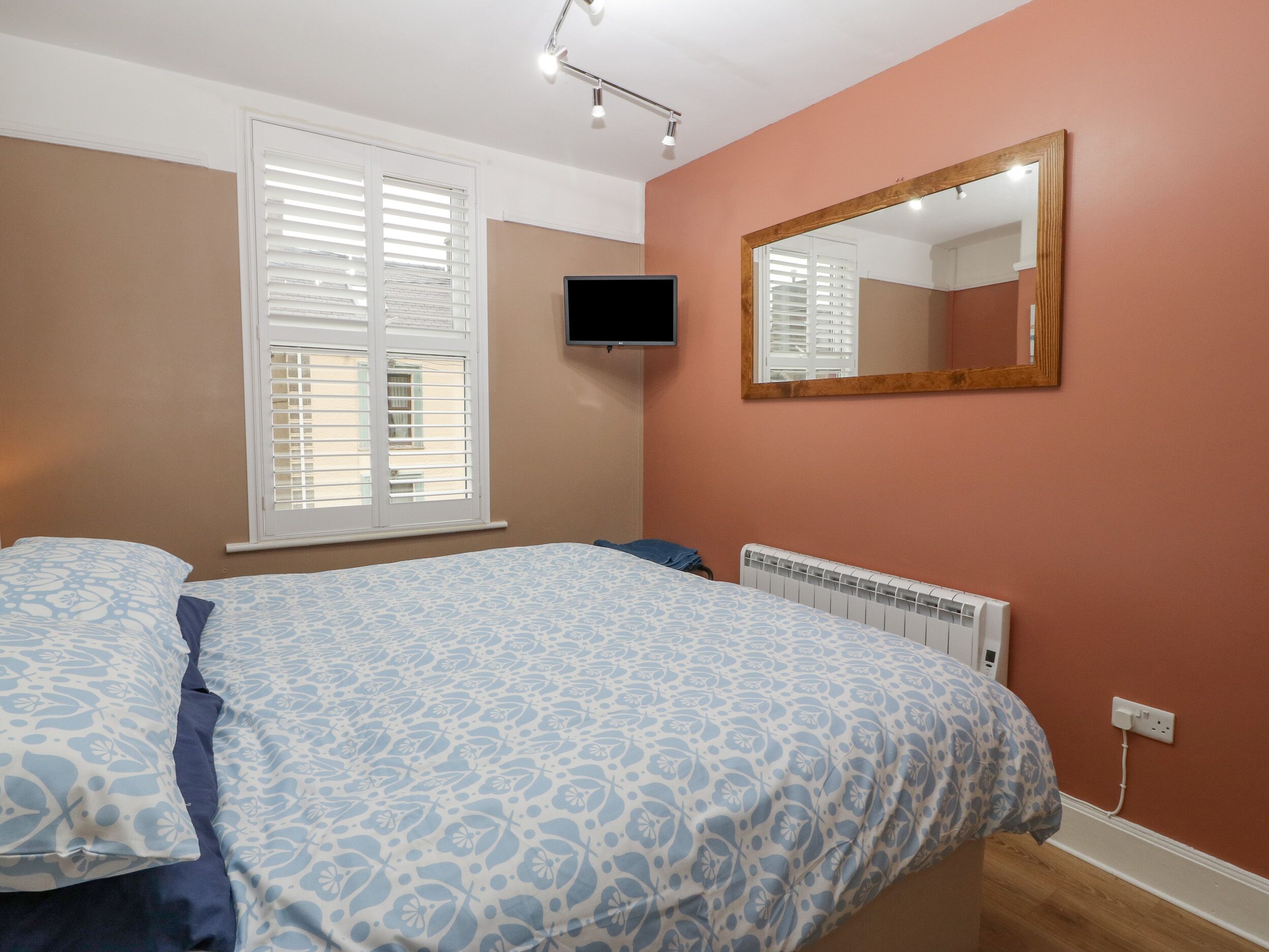 A bedroom with a bed, a television, a window with blinds, and a wall-mounted mirror.
