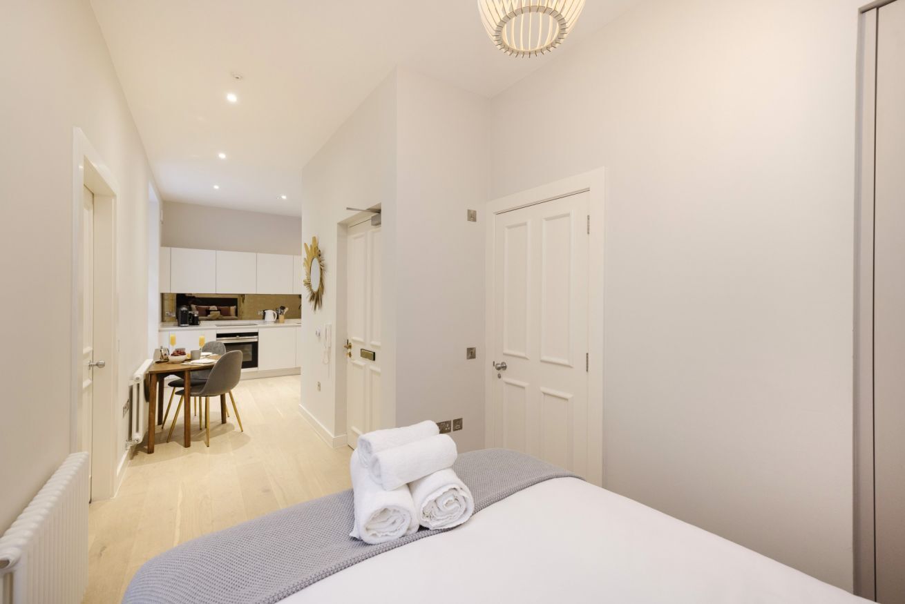 A modern hotel room with a neatly made bed, a dining area with a table and chairs, and a kitchen in the background.