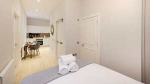 A modern hotel room with a neatly made bed, a dining area with a table and chairs, and a kitchen in the background.