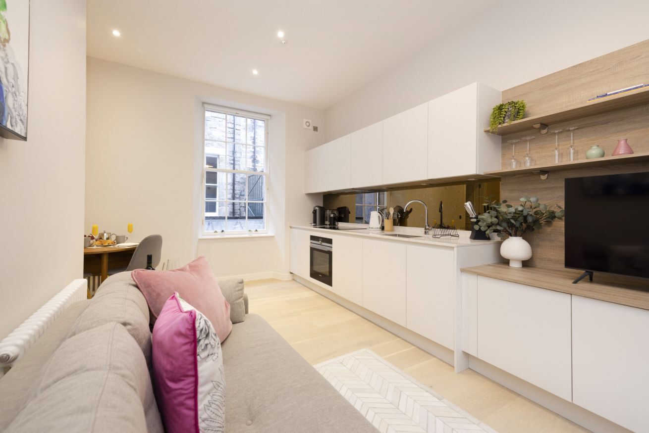 A modern kitchen with white cabinets, a wooden countertop, and a built-in oven. There is a dining area with a table and chairs, and a large window providing natural light.