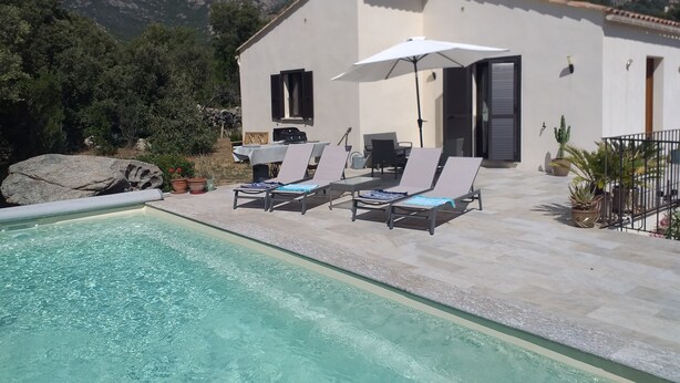 Ein Poolbereich mit Liegestühlen und Sonnenschirm, ein weißes Gebäude mit Blick auf Berge und ein klarer Himmel.