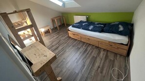 Une chambre située dans un espace de type « loft », avec une lucarne, un plancher en bois, un lit avec une literie bleue et une petite table en bois.