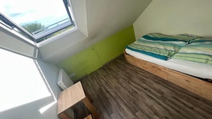 Une petite pièce aux murs inclinés, dotée d’un plancher en bois, d’un lit recouvert d’une couverture à rayures et d’une fenêtre donnant sur la verdure.