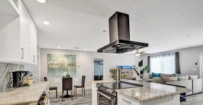 A modern kitchen with a central island, a dining area with chairs, and a living space with a sofa and a large window.