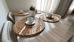 Un coin repas avec une table en bois, des sets de table en fibres tressées, des chaises blanches et une plante en pot.
