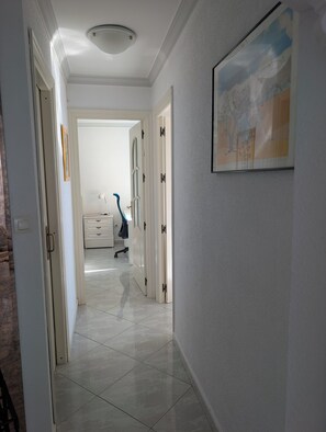 A hallway with a tiled floor, a painting on the wall, and a door leading to a room with a desk and chair.