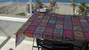 A balcony with a table covered in a colorful tablecloth, overlooking a beach with palm trees and a clear blue sky.