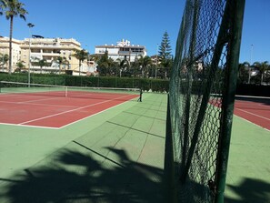A tennis court with a chain-link fence, surrounded by buildings and palm trees.