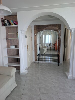 A spacious living area with a white sofa, a bookshelf, and a clear view into a bedroom through an arched doorway.