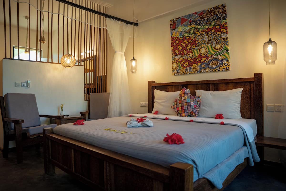 A neatly made bed with a colorful pillow, a bedside table, a chair, and a wall with a patterned artwork.