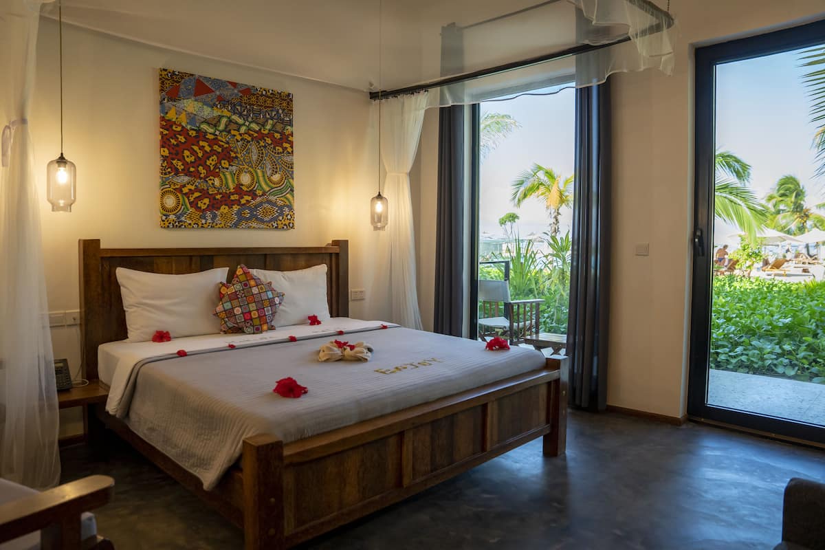 A bedroom with a wooden bed, a colorful artwork on the wall, and a view of a tropical garden through the sliding glass door.