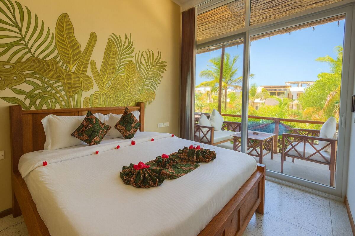 A hotel room with a large bed, a balcony with outdoor seating, and a view of greenery and buildings.
