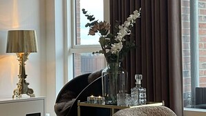 Moderni hotellihuone, jossa on suuri ikkuna, musta tuoli, yöpöytä lampun kanssa ja maljakko kukkineen.