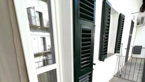Un balcon doté d’une porte-fenêtre coulissante et de volets verts, d’un sol en béton et d’un mur blanc.