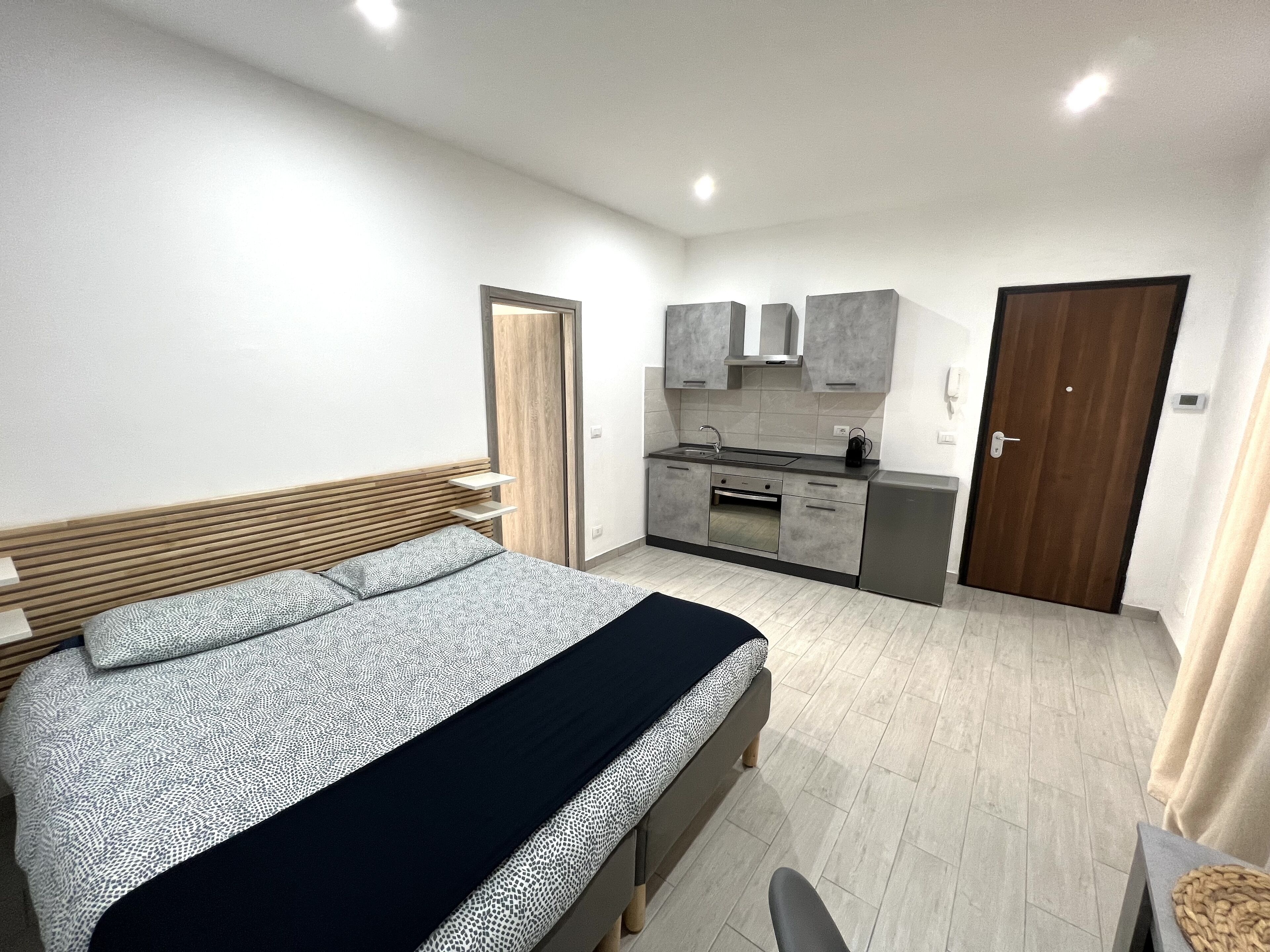 A bedroom with a bed, bedside table, and a small desk with a chair. There is a kitchenette with a sink, stove, and cabinets.