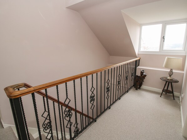 A staircase with a wooden handrail and black metal balusters, carpeted floor, a small table with a lamp, and a window with blinds.
