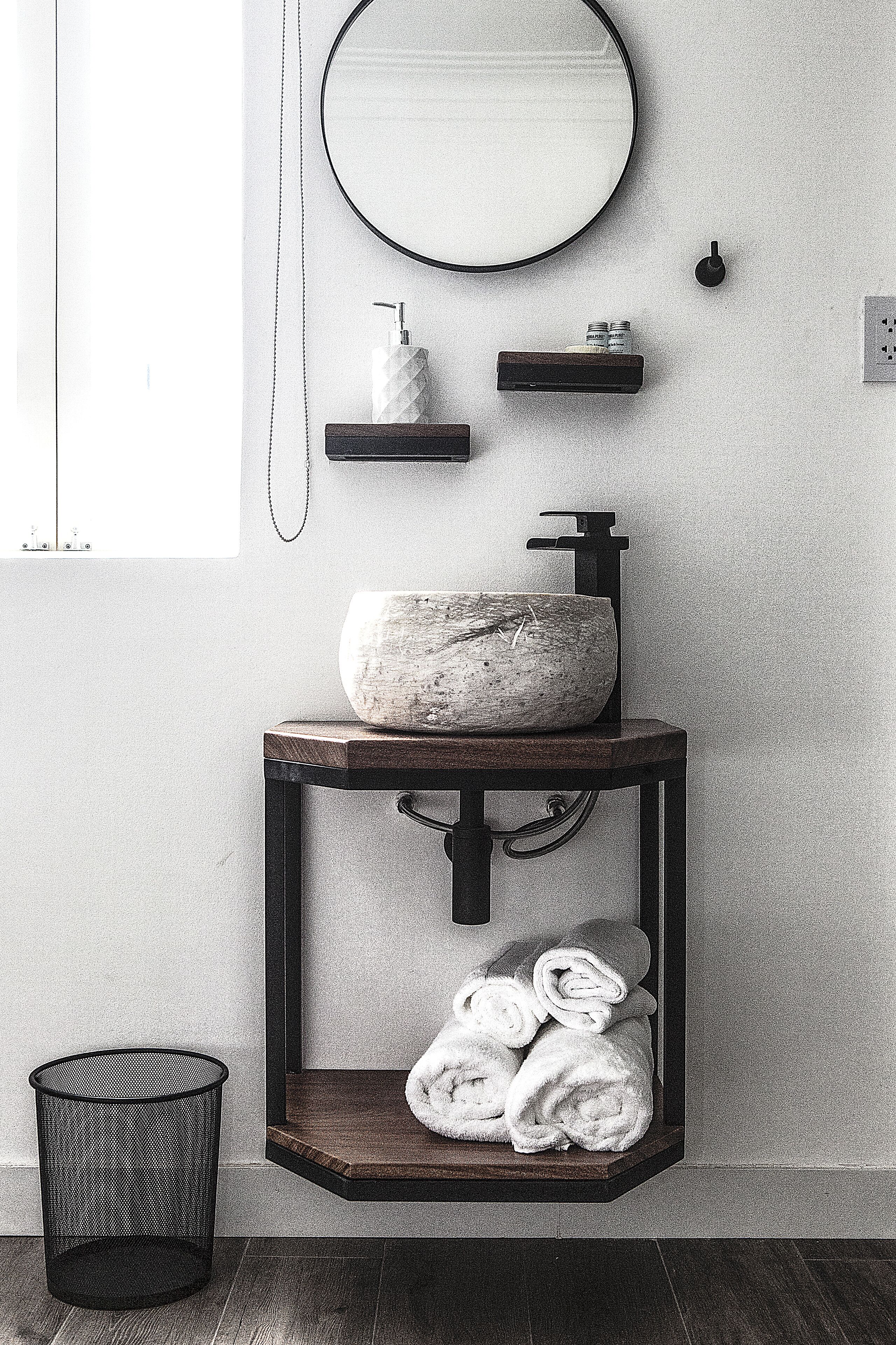 A bathroom with a round mirror, a stone sink, a towel rack, and a black trash can.