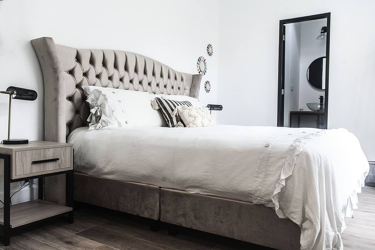 Een slaapkamer met een gestoffeerd hoofdeinde, een nachtkastje met een lamp en een spiegel.