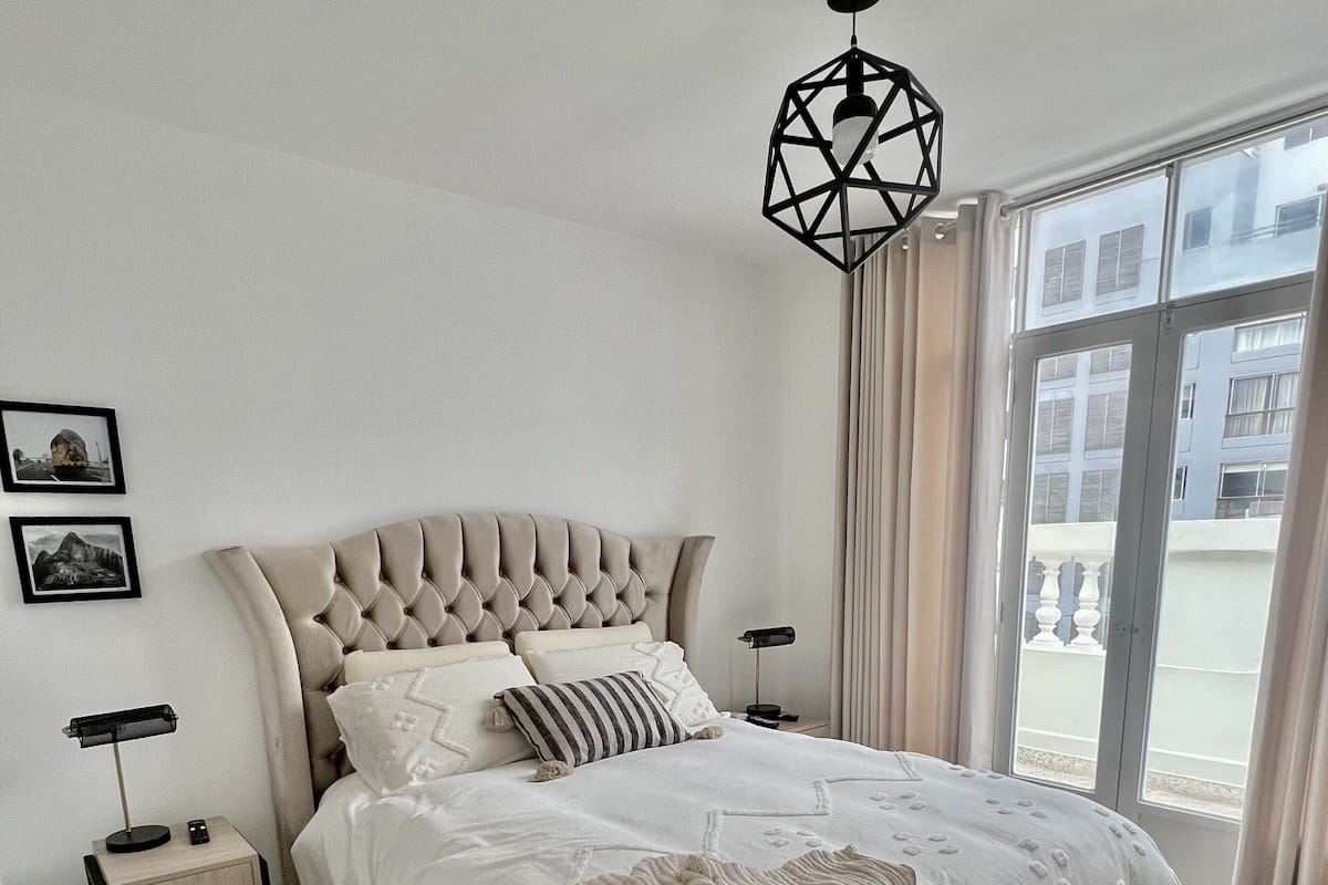 Een moderne slaapkamer met een gestoffeerd hoofdeinde, een groot raam met gordijnen, een nachtkastje met een lamp en een decoratieve wandversiering.