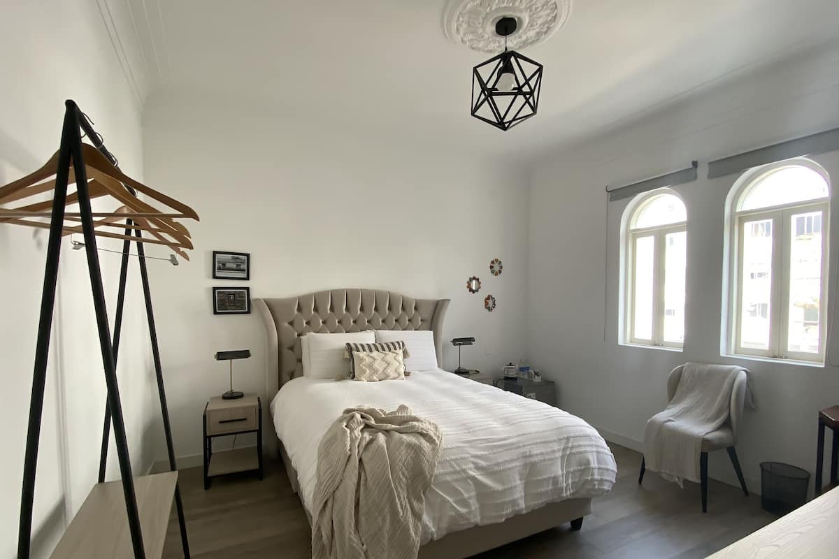 Een slaapkamer met een groot bed, een houten kledingrek, een nachtkastje met een lamp en twee ramen.