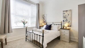 A bedroom with a bed, bedside tables, a nightstand, a window with blinds, and a painting on the wall.